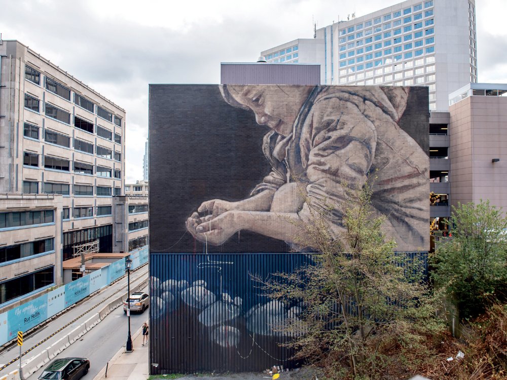 A large mural on a building wall depicts a child in a hoodie, intently focused on their hands. The scene conveys a quiet, contemplative mood. Urban setting with cars and trees in the foreground.