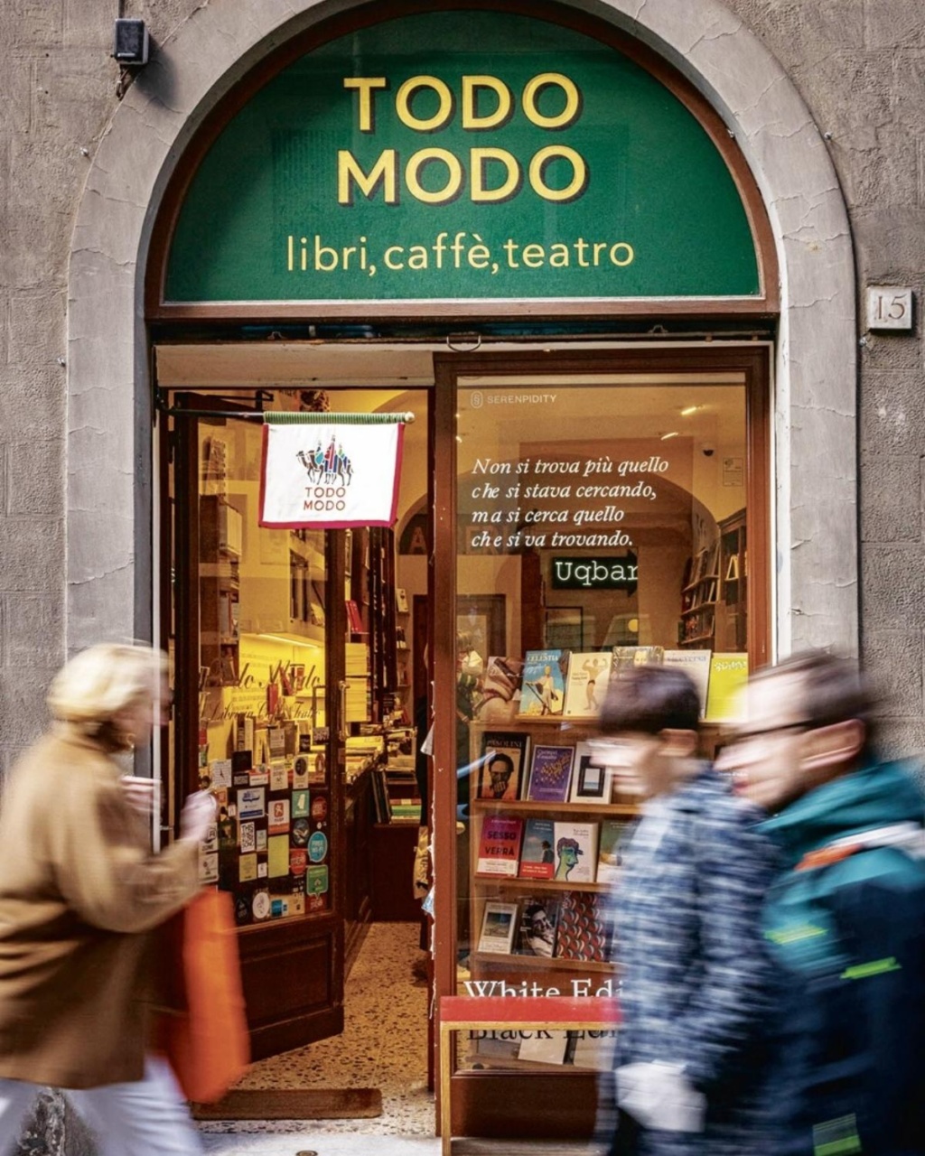 A cozy bookstore entrance with a green sign saying "Todo Modo libri, caffè, teatro." Blurred pedestrians walk past, creating a lively, bustling atmosphere.