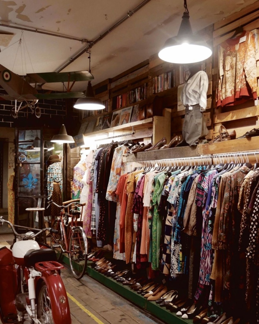 Vintage clothing store with colorful dresses hanging on racks, old bicycles on the left, and a model airplane above, creating a nostalgic ambiance.