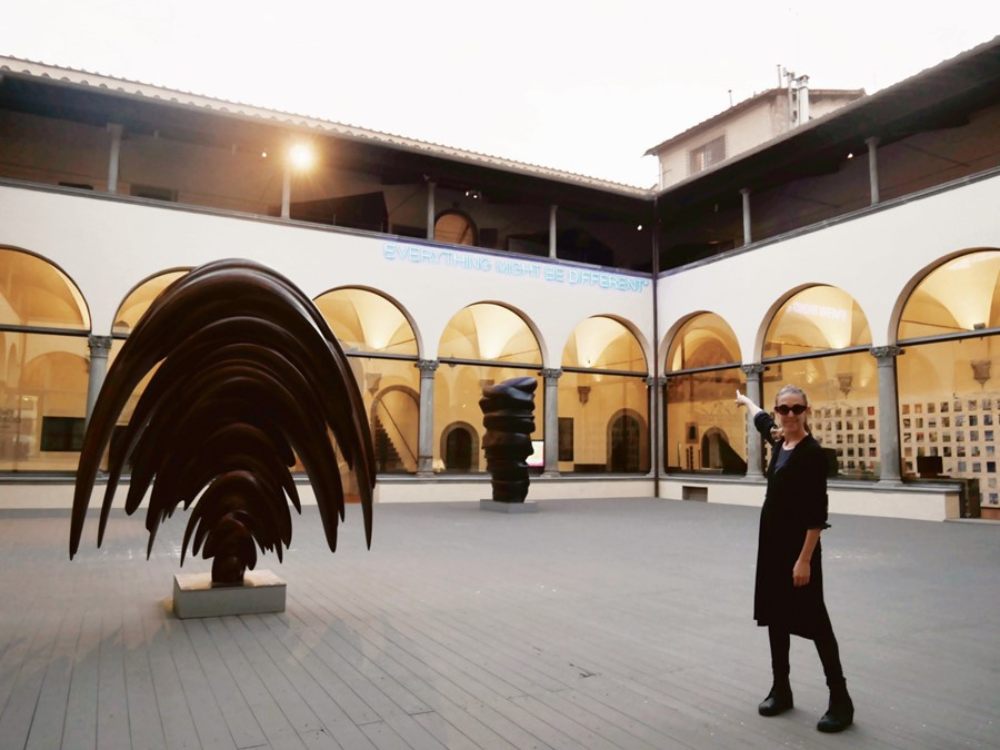 A person in black clothing gestures towards modern sculptures in an open, arched courtyard of an art museum. The setting is calm and contemplative.
