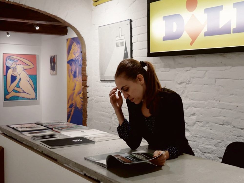 Woman with ponytail reading at a gallery counter, surrounded by modern art on white walls. The scene feels focused and contemplative.