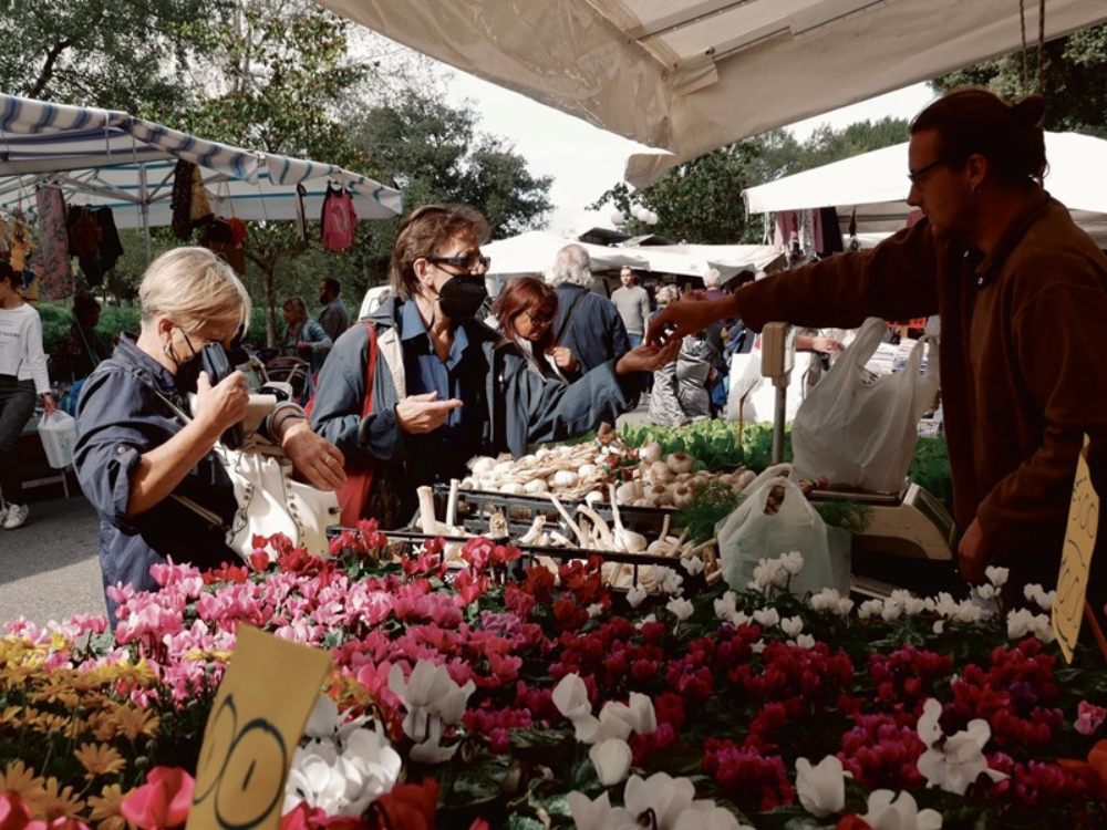 A bustling outdoor market with colorful flowers in the foreground. People in masks are buying produce, creating a lively and vibrant atmosphere.