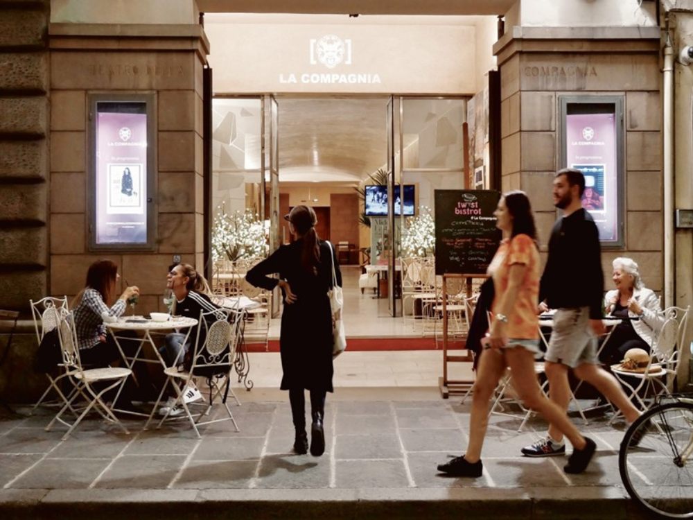 A cozy café entrance with outdoor seating on a cobblestone street. Two people chat at a table while others walk by, creating a lively and inviting atmosphere.