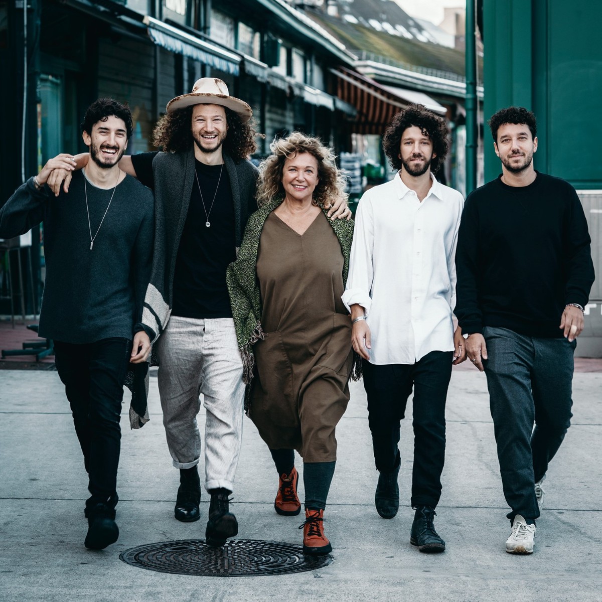 A group of five people, three men and two women, walk happily down a street. They are closely positioned, smiling and casual, conveying camaraderie. The setting is urban with buildings in the background.