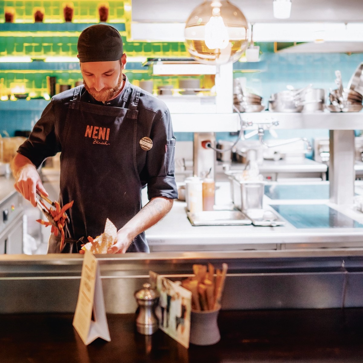 Chef in a black uniform prepares food in a brightly lit kitchen with a warm ambiance. Kitchen utensils and shelves with pots are visible in the background.