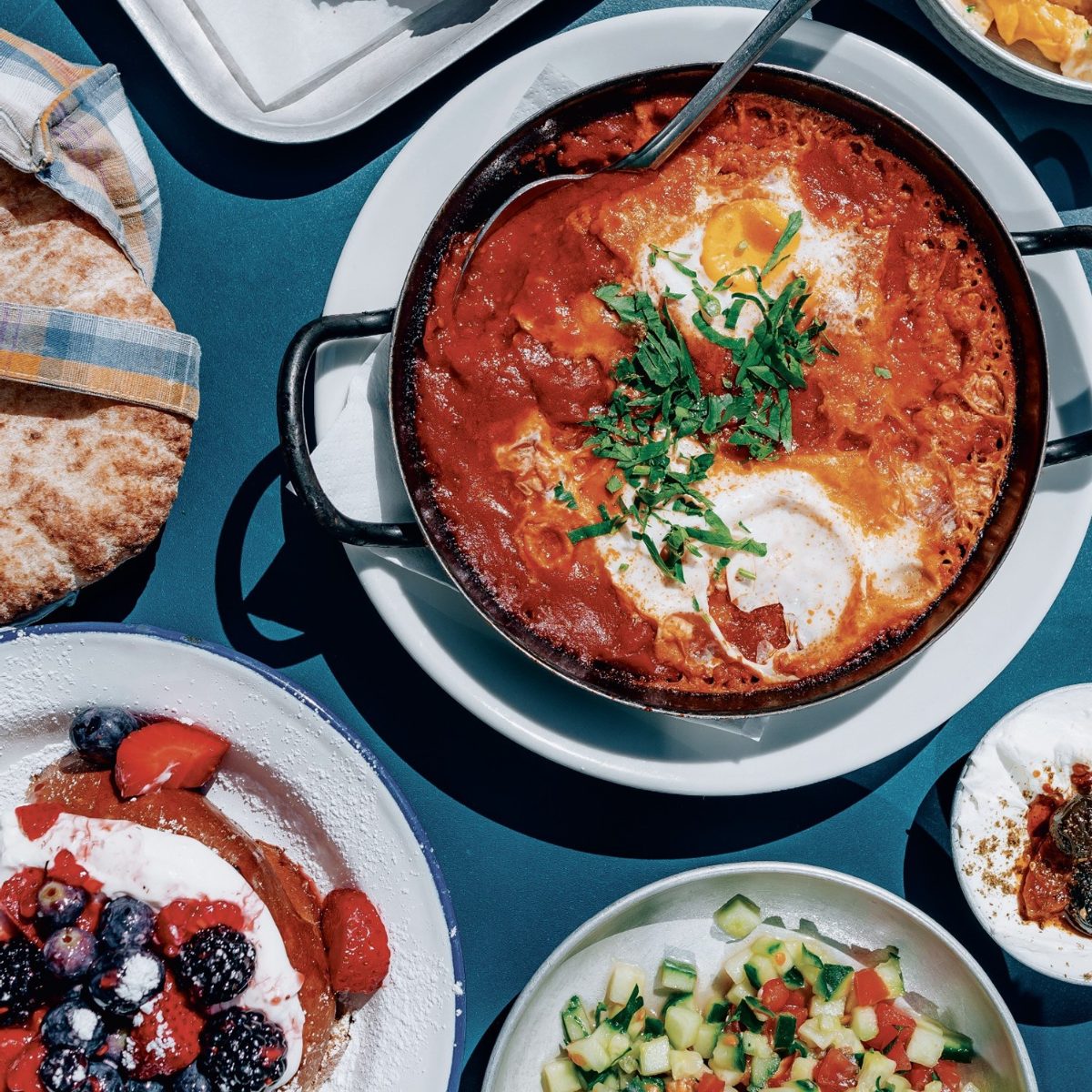 A colorful spread of Middle Eastern dishes on a blue table. A skillet of shakshuka with eggs and herbs, pita bread, berries with cream, and a cucumber salad.