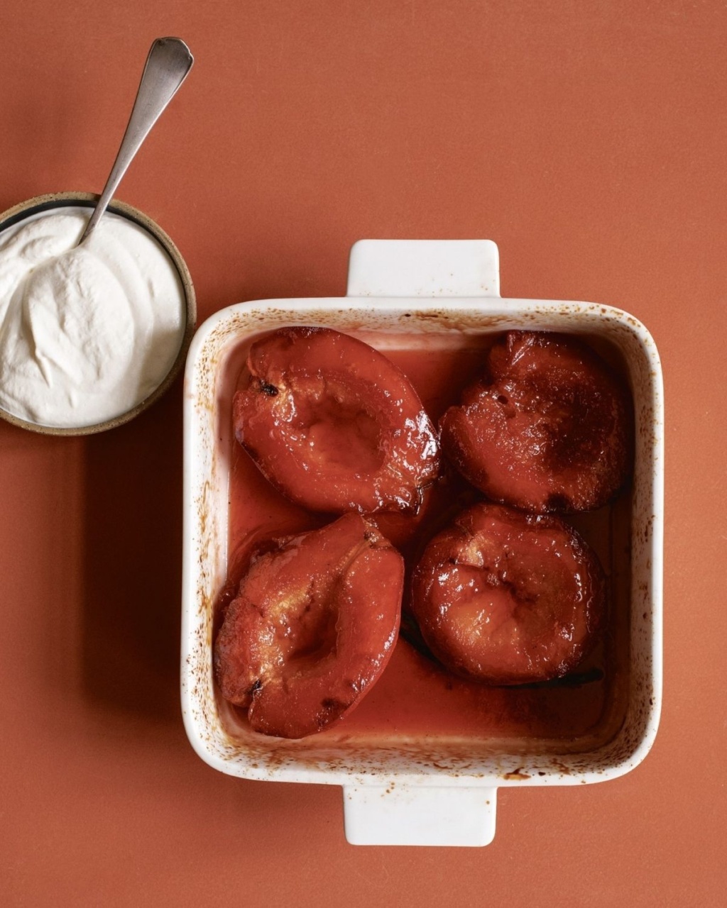 Roasted quince halves in a white baking dish with syrup, next to a small bowl of creamy white yogurt, set on a warm terracotta background. Cozy and inviting.