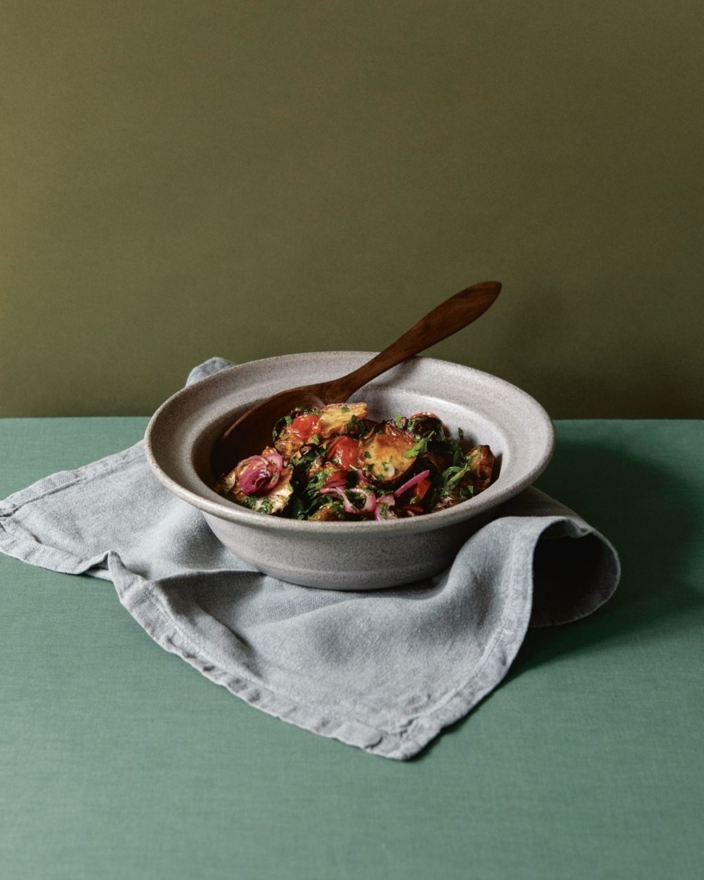 Ceramic bowl with roasted vegetables and herbs, placed on a gray napkin over a green textured surface. A wooden spoon rests inside, against an olive backdrop.