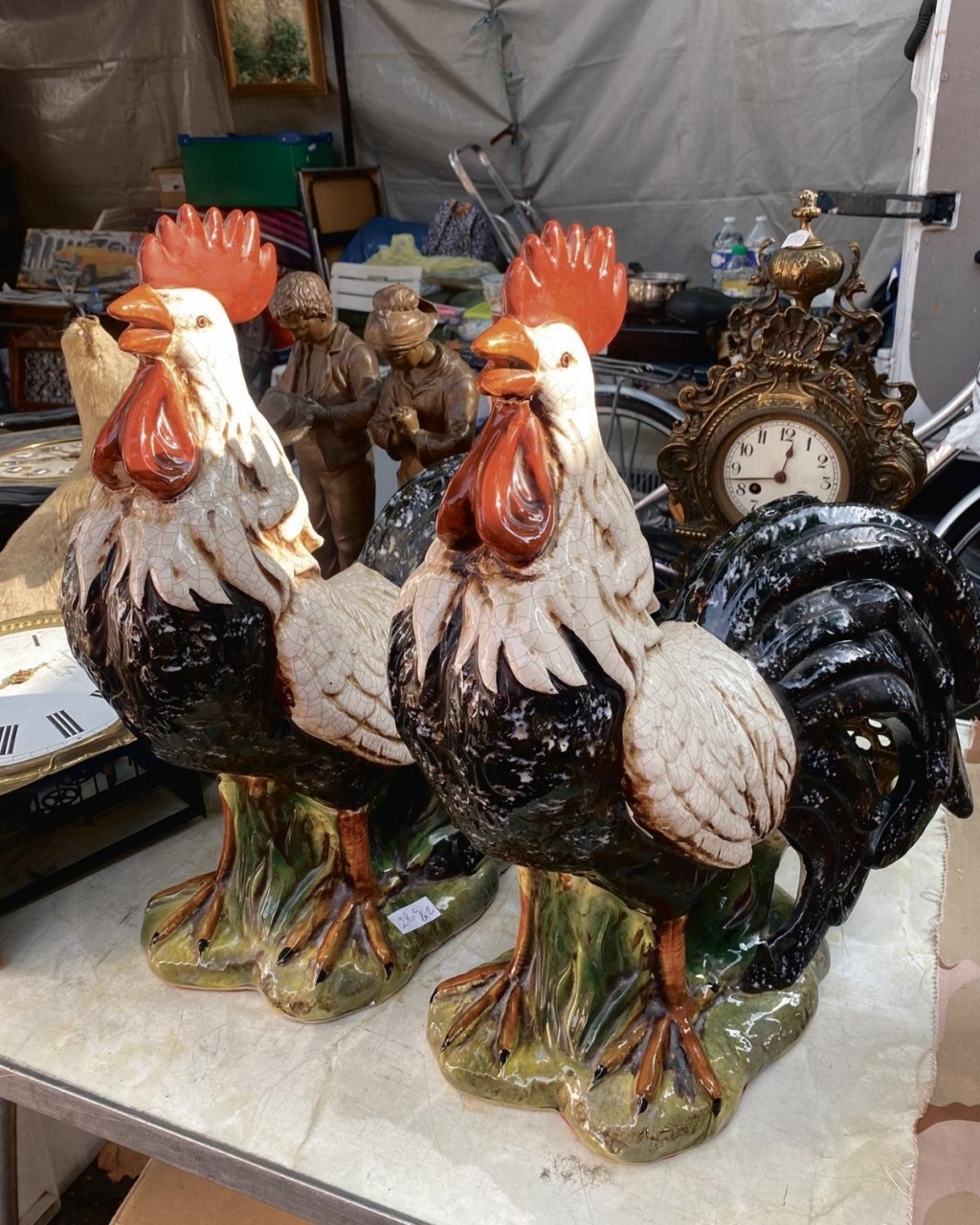 Two ornate ceramic rooster statues with vivid red combs and detailed feathers stand on a table at a flea market, surrounded by vintage items.