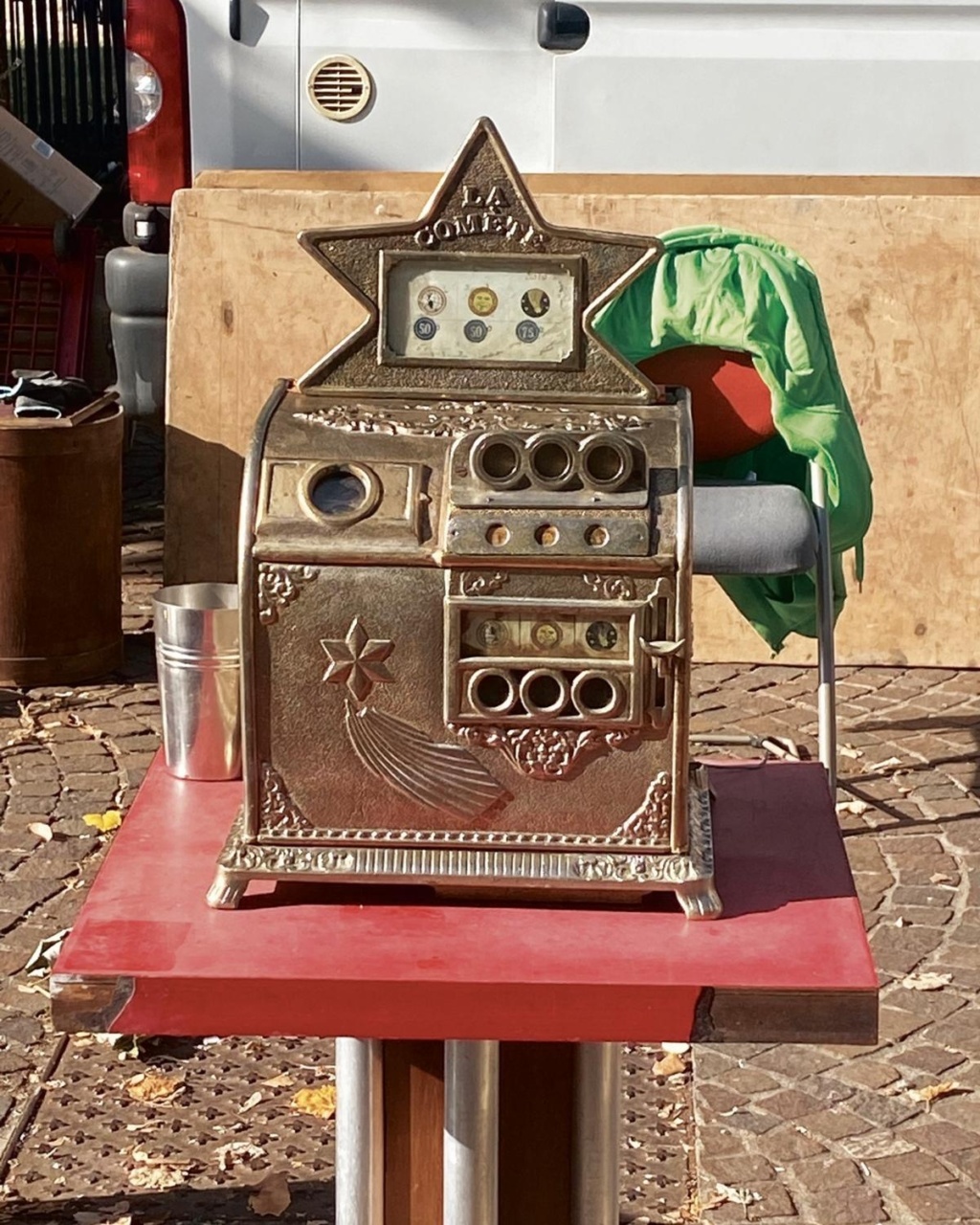 Vintage silver cash register with a star-shaped top labeled "La Cometa" on a red table. Background shows a pavement and white van. Retro vibe.