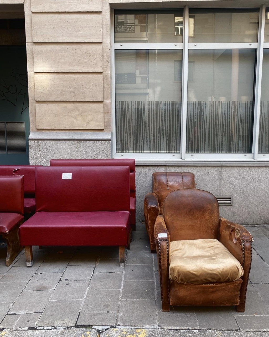 Old red and brown leather sofas stand side by side on a city sidewalk against a building wall and windows, creating an abandoned, nostalgic vibe.
