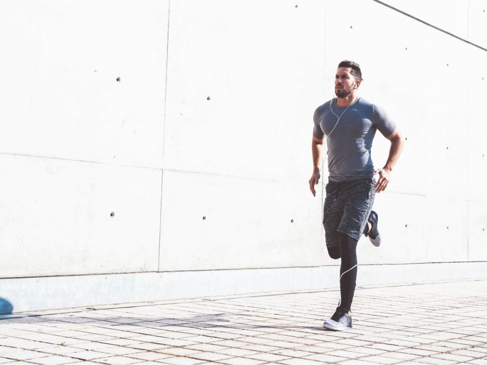 A man in athletic wear runs along a sunlit concrete path, earphones in, exuding focus and energy. A minimalist, urban backdrop enhances the scene.