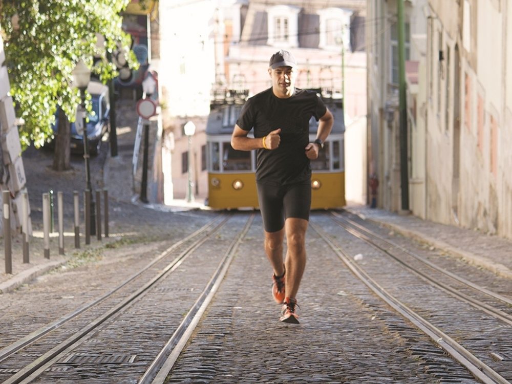 A man in athletic wear jogs uphill on a cobblestone street with tram tracks. A yellow tram approaches from behind, set in a sunny urban scene.