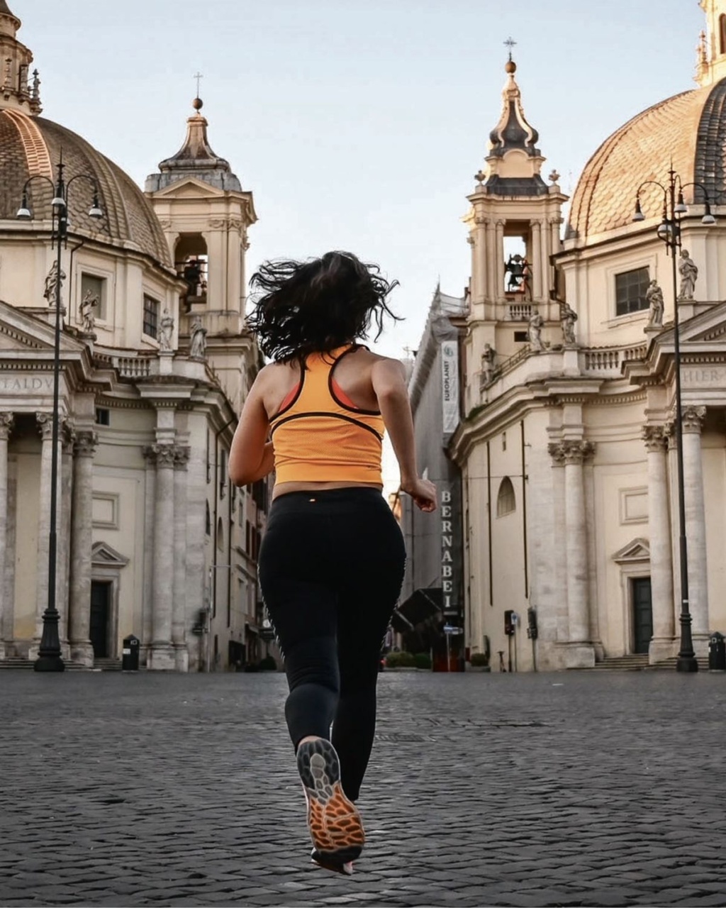 A woman in athletic gear runs through an empty cobblestone plaza flanked by ornate, domed buildings under a clear sky, evoking energy and freedom.