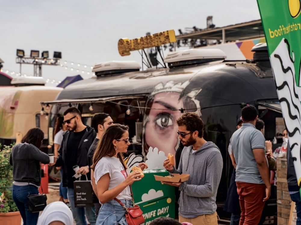 People enjoying food at an outdoor market with a food truck in the background. The truck features vibrant graffiti art. The mood is lively and social.