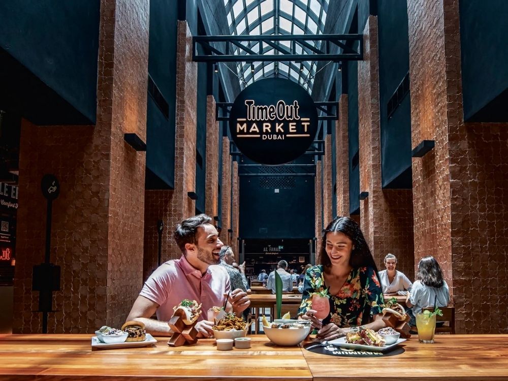 A smiling couple sits at a wooden table, enjoying food and drinks at Time Out Market Dubai. The setting is lively, with a modern, stylish ambiance.