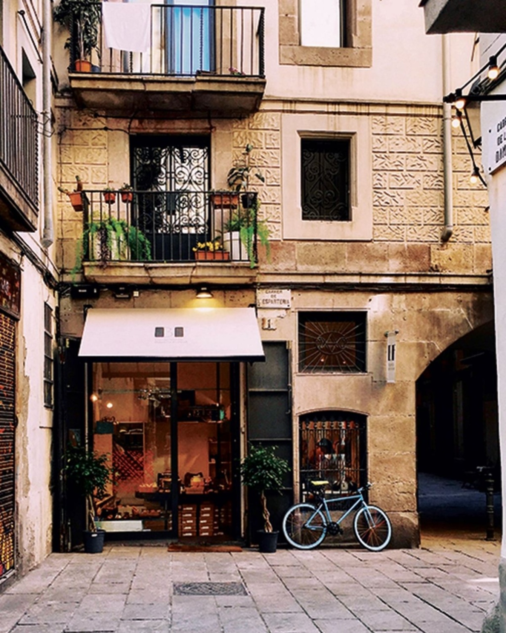 A quaint street scene features a cozy shop with a white awning, flanked by potted plants and a blue bicycle parked nearby. The building's facade is adorned with rustic balconies and greenery, evoking a charming, relaxed mood.