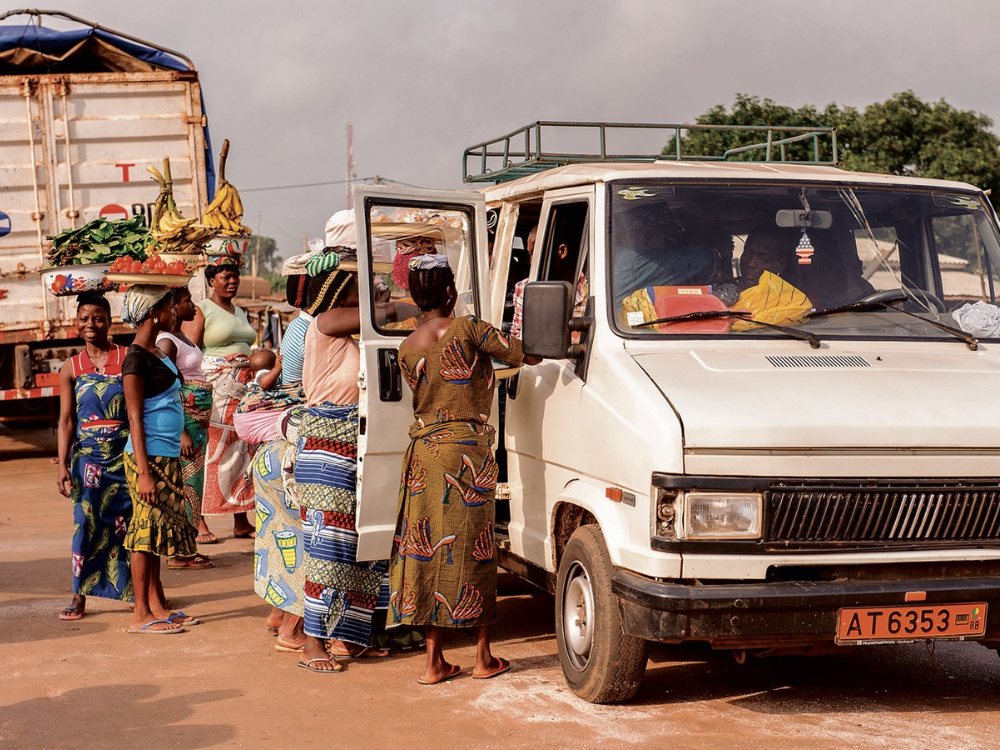 A group of women, some balancing goods on their heads, gather around a parked white van on a dusty road. The scene feels vibrant and busy.