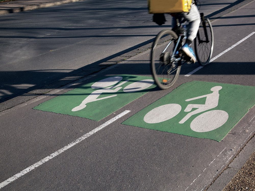 A cyclist with a yellow backpack rides on a bike lane marked with green symbols of bicycles. Shadows and motion blur suggest a sunny day and movement.