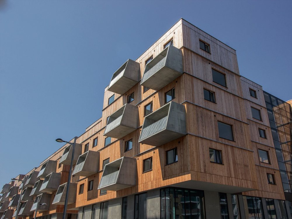 Modern building with wooden façade and protruding boxy balconies, set under a clear blue sky. The structure conveys a geometric, contemporary feel.
