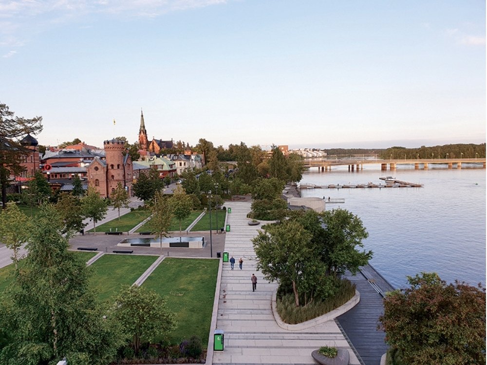 A riverside park with manicured lawns, trees, and wide walkways. A historic brick building and a church spire rise in the town, with a serene river view.