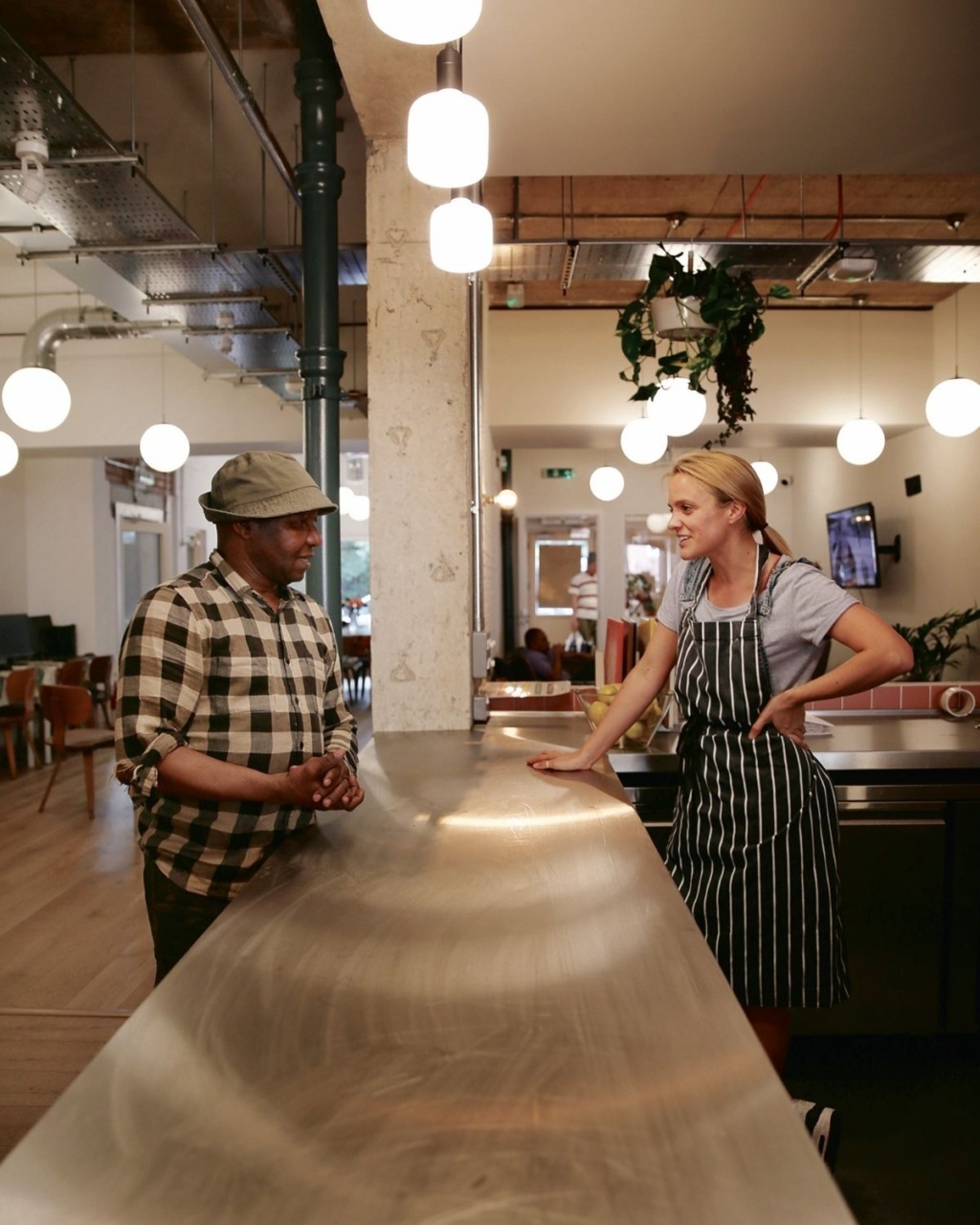 A man in a plaid shirt talks to a woman in a striped apron behind a counter in a cosy café, under warm hanging lights.
