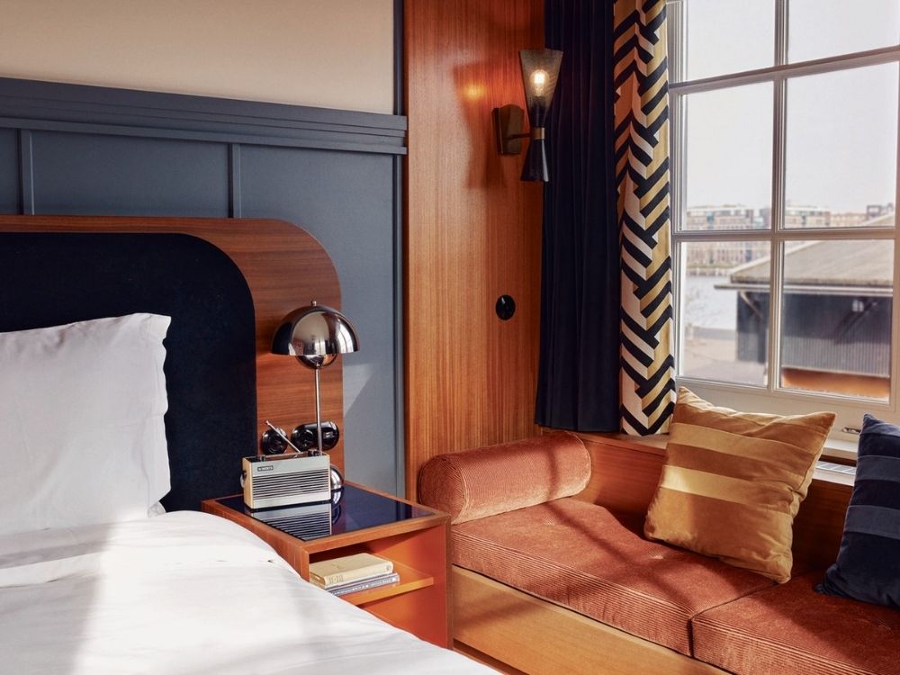 Cozy hotel room with wood paneling and a large window. A bed with white linens, a retro radio, a stylish lamp, and a velvet sofa with striped pillows create an inviting vibe.