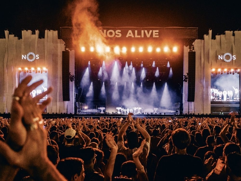 Crowd cheering at a nighttime outdoor concert under "NOS Alive" sign. Bright stage lights, enthusiastic atmosphere, and large screens on each side display performers.