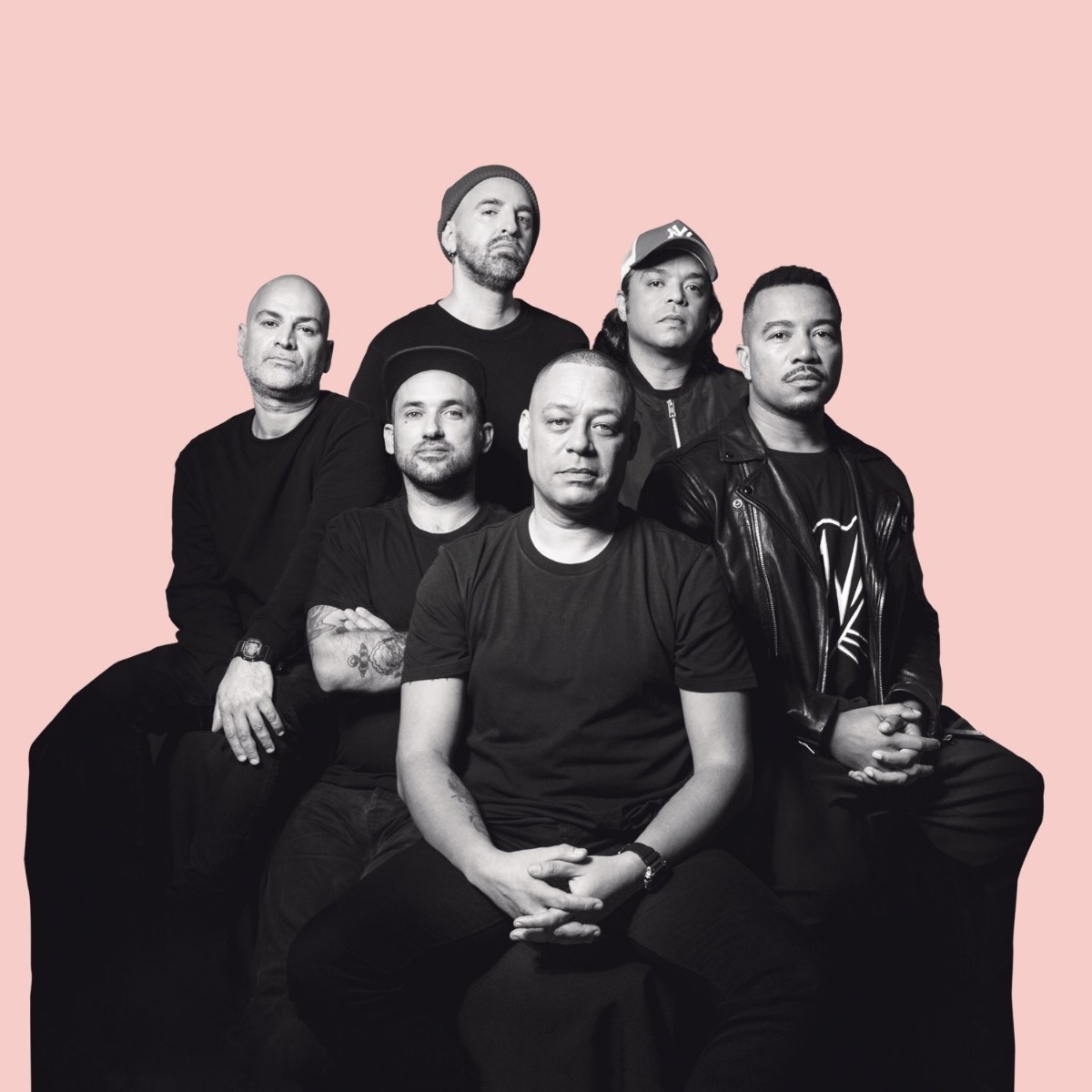 Six men sit and stand arranged in two rows against a pink background, wearing casual black clothing, exuding a calm and confident demeanor.