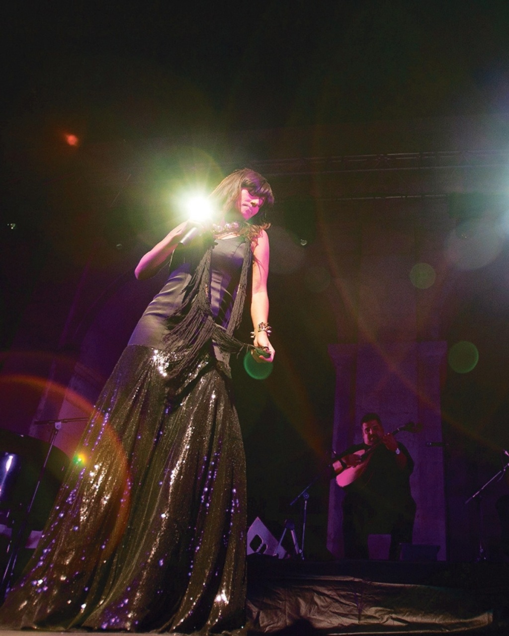 A woman in a glittering black dress sings passionately on stage, bathed in colorful spotlights. A musician plays guitar in the background, creating a vibrant ambiance.