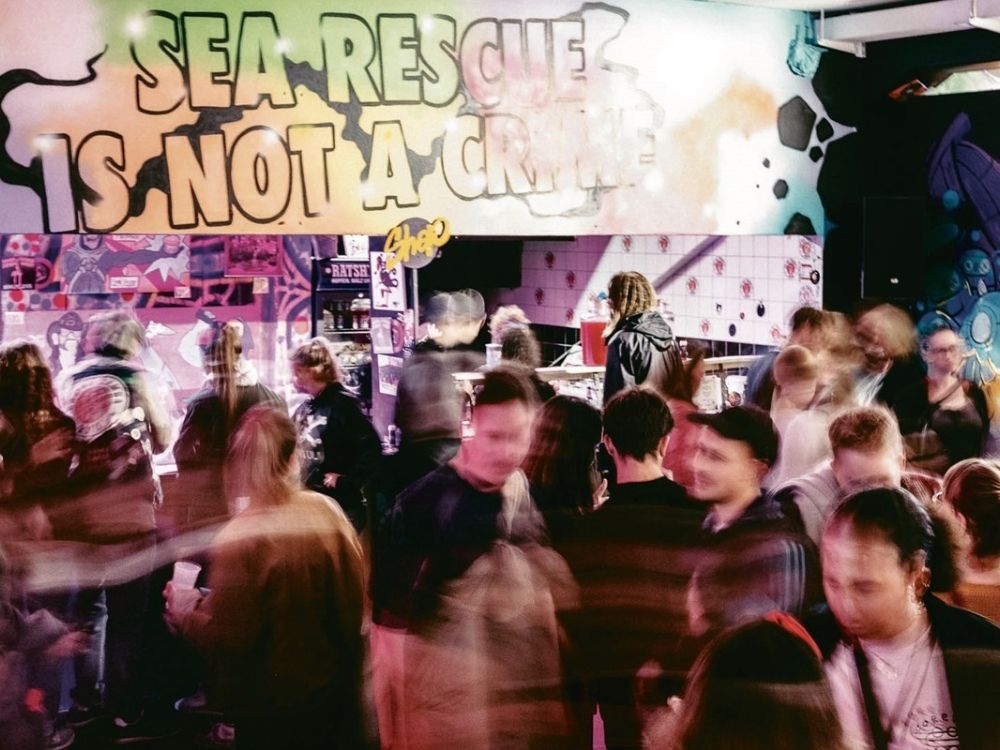 A lively crowd in a vibrant room with graffiti stating "Sea Rescue Is Not A Crime."