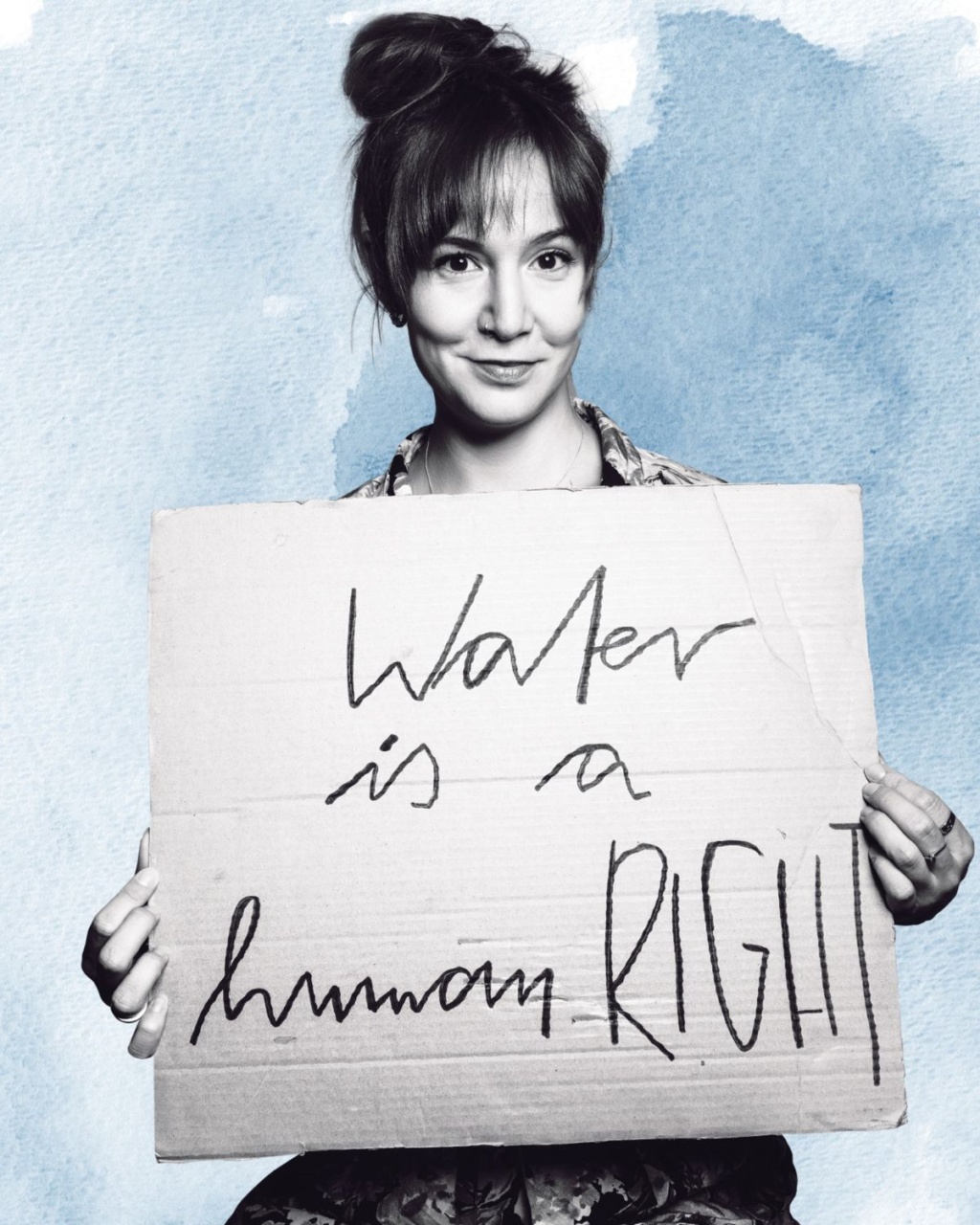 A person with a topknot hairstyle holds a cardboard sign reading "Water is a human RIGHT" against a blue watercolor background.