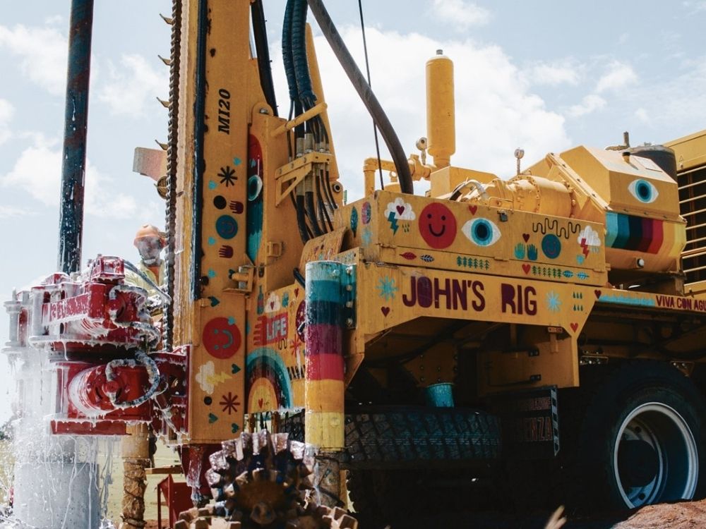 Colorful, decorated drilling rig labeled "John's Rig," with vibrant patterns and happy motifs, sprays water under a sunny sky.