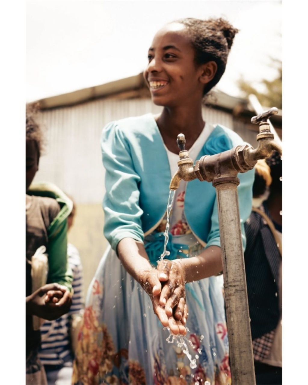 A young girl joyfully washes her hands under a tap, wearing a bright blue jacket and floral dress.