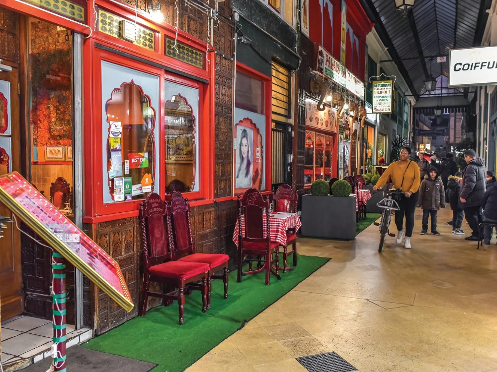 A cozy street with a restaurant featuring red decor and checkered tablecloths on the left. People walk past, creating a lively, inviting atmosphere.