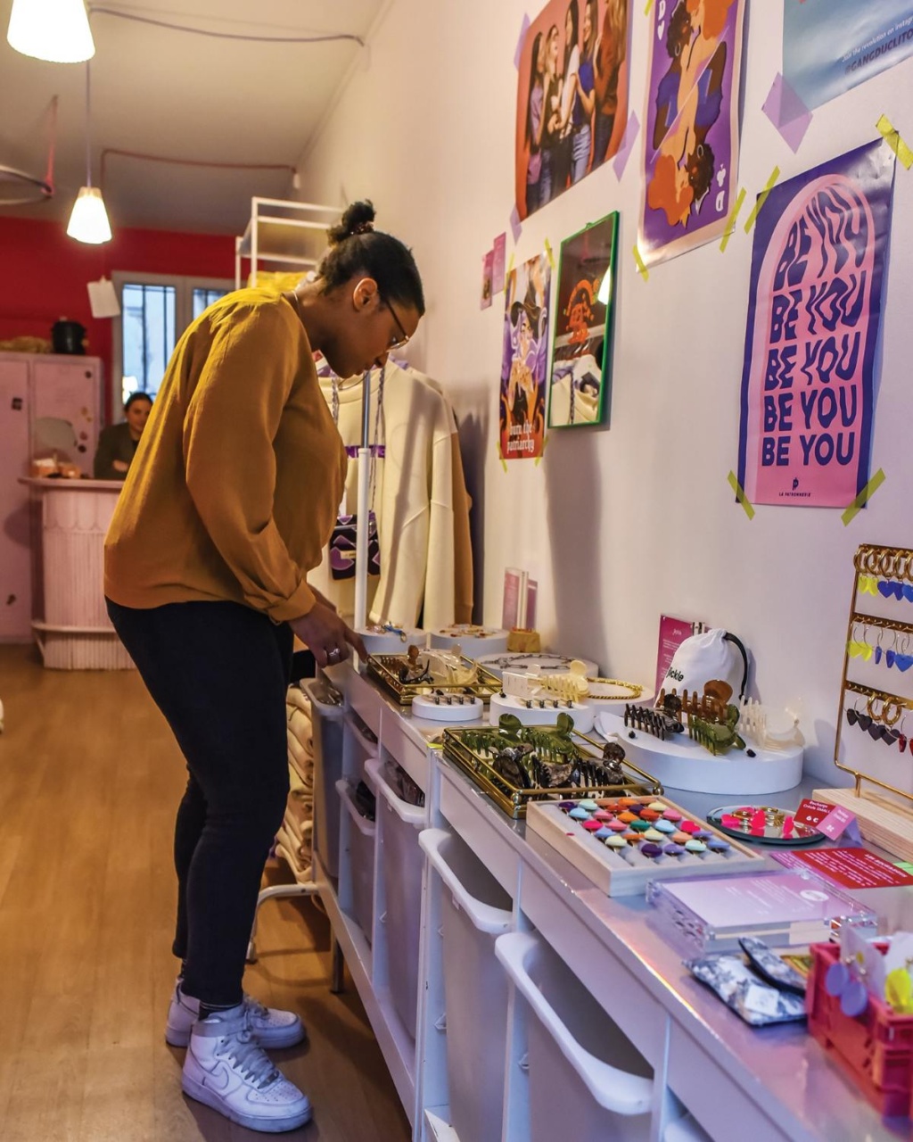 A person in a mustard sweater examines jewelry in a boutique with colorful art posters on the wall, creating a cozy and creative atmosphere.