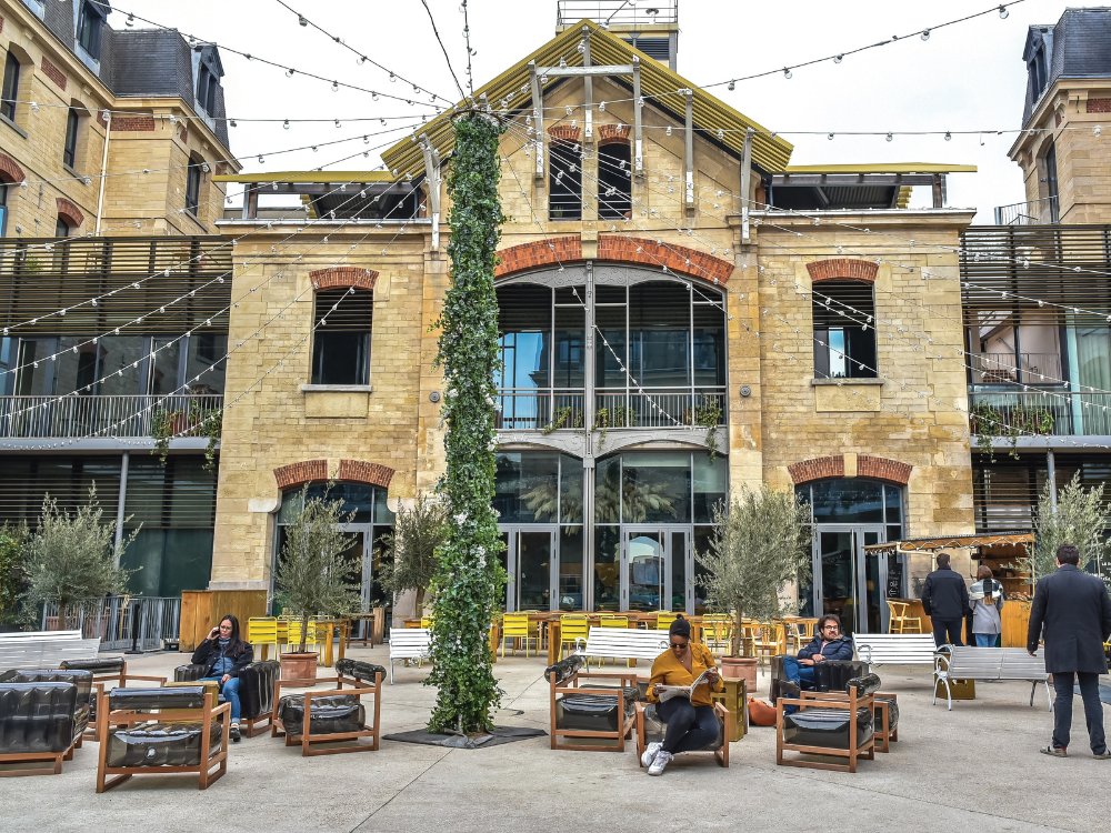A historical stone building with arched windows serves as a backdrop for an outdoor seating area. People are relaxing on modern wooden furniture under string lights, creating a casual, inviting atmosphere.