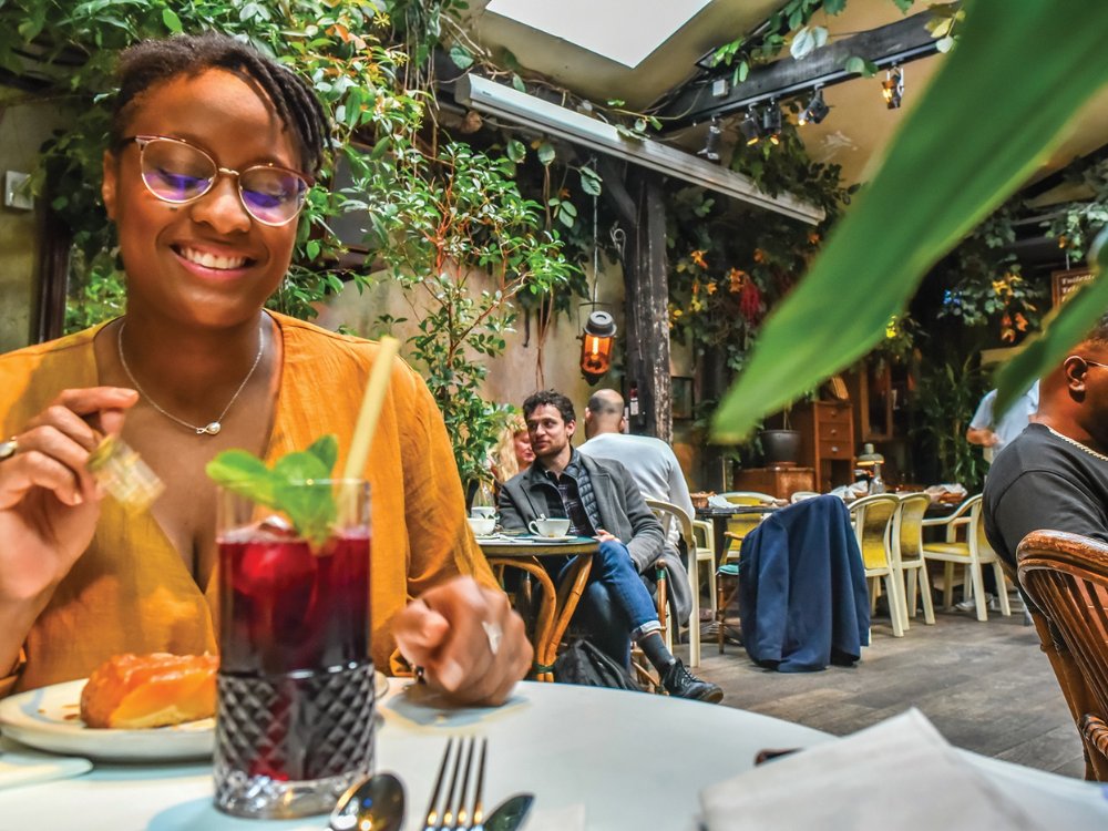 Smiling woman in a cozy, plant-filled café with a stylish drink and dessert. Warm, inviting atmosphere with people chatting in the background.