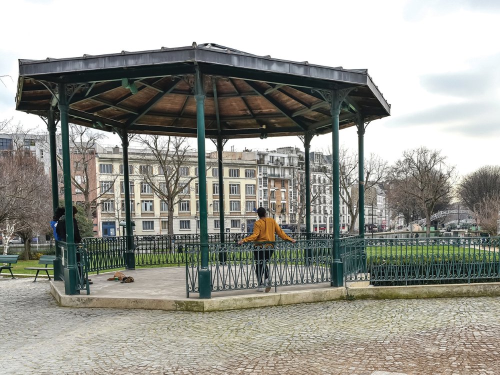 A person in a mustard coat stands on a gazebo in a park, surrounded by bare trees and buildings. The setting feels peaceful and contemplative.
