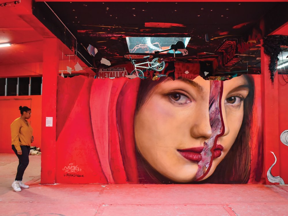 A vibrant mural features a woman's face on a red wall, her expression serene yet mysterious. A person walks nearby, adding a sense of scale.