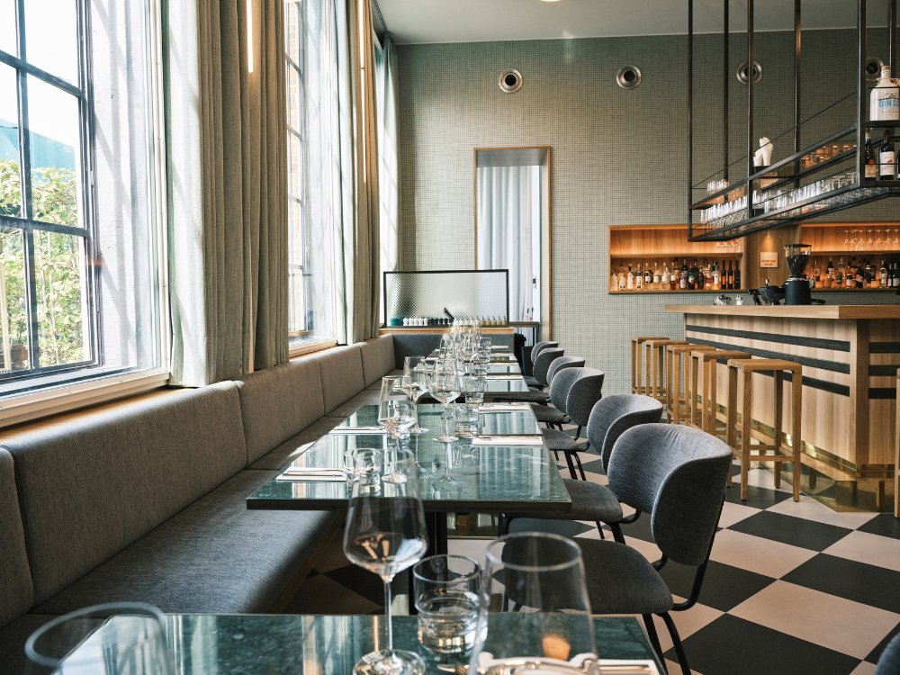 Modern restaurant interior featuring a long seating area, glass tables, and a stylish bar, illuminated by natural light from large windows.