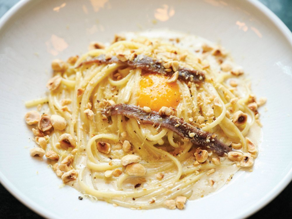 A bowl of spaghetti topped with a runny egg yolk, crispy anchovies, and crushed hazelnuts, garnished with a light creamy sauce.