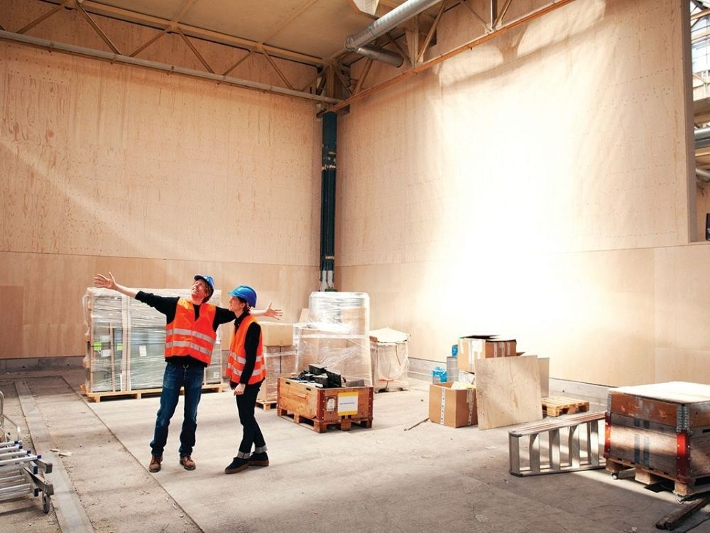 Two people in hard hats and orange safety vests stand in a large, empty warehouse with wooden walls.