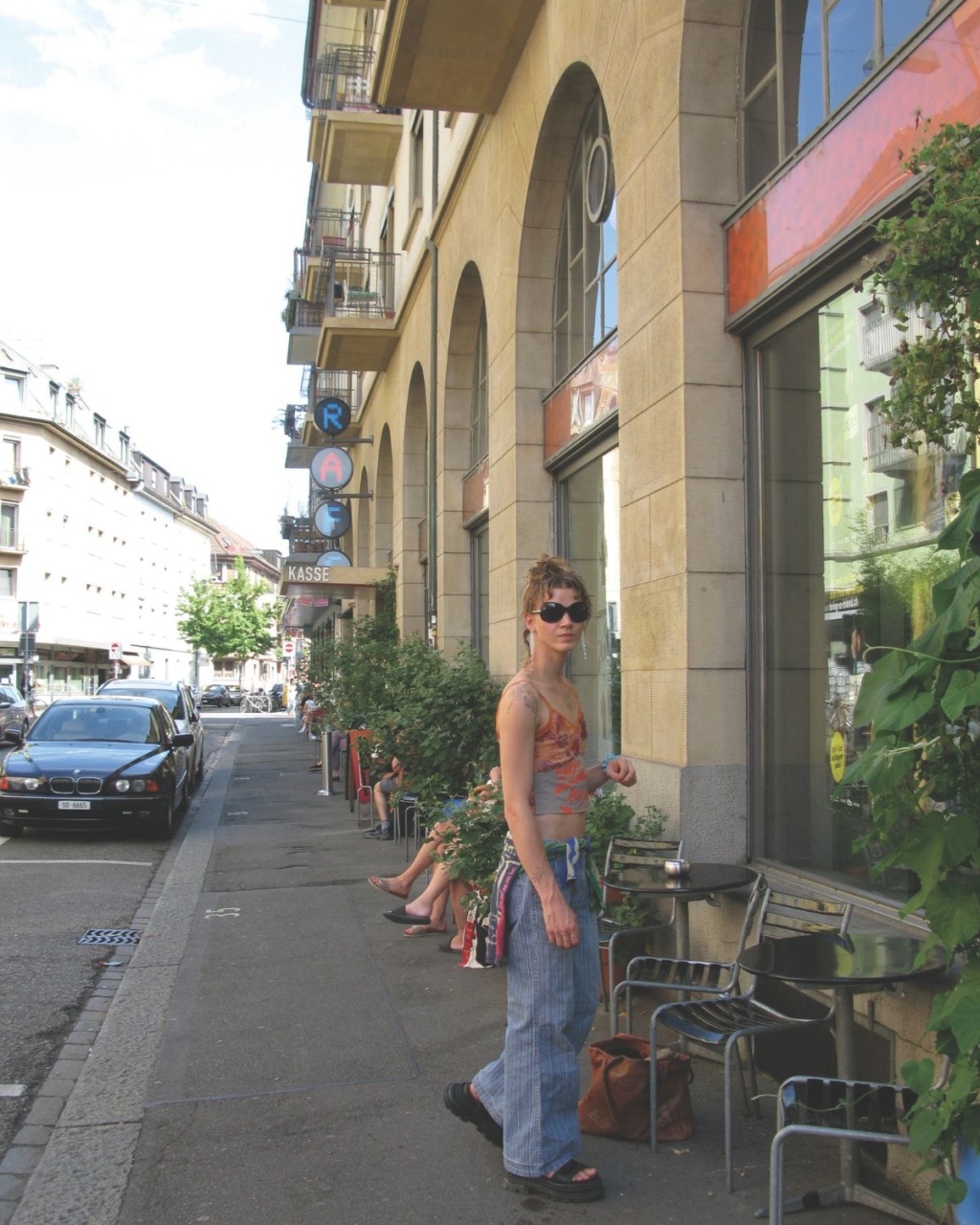 Luca stands on a sunlit sidewalk beside an arched building. Cafes and plants line the street, with city life in the background.