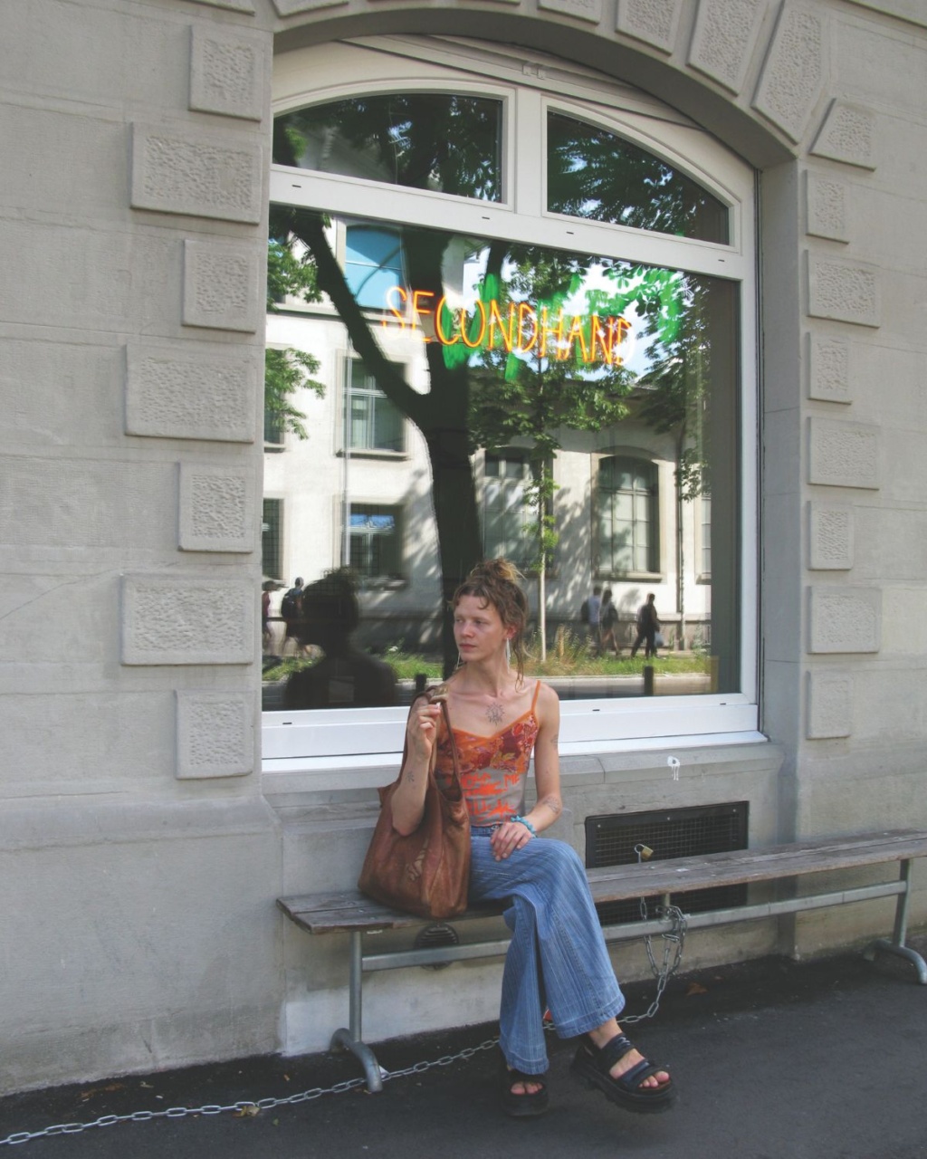 Luca with a brown bag sits on a bench outside a building. She's in casual clothes, and the window above her reflects trees and the word "Secondhand."