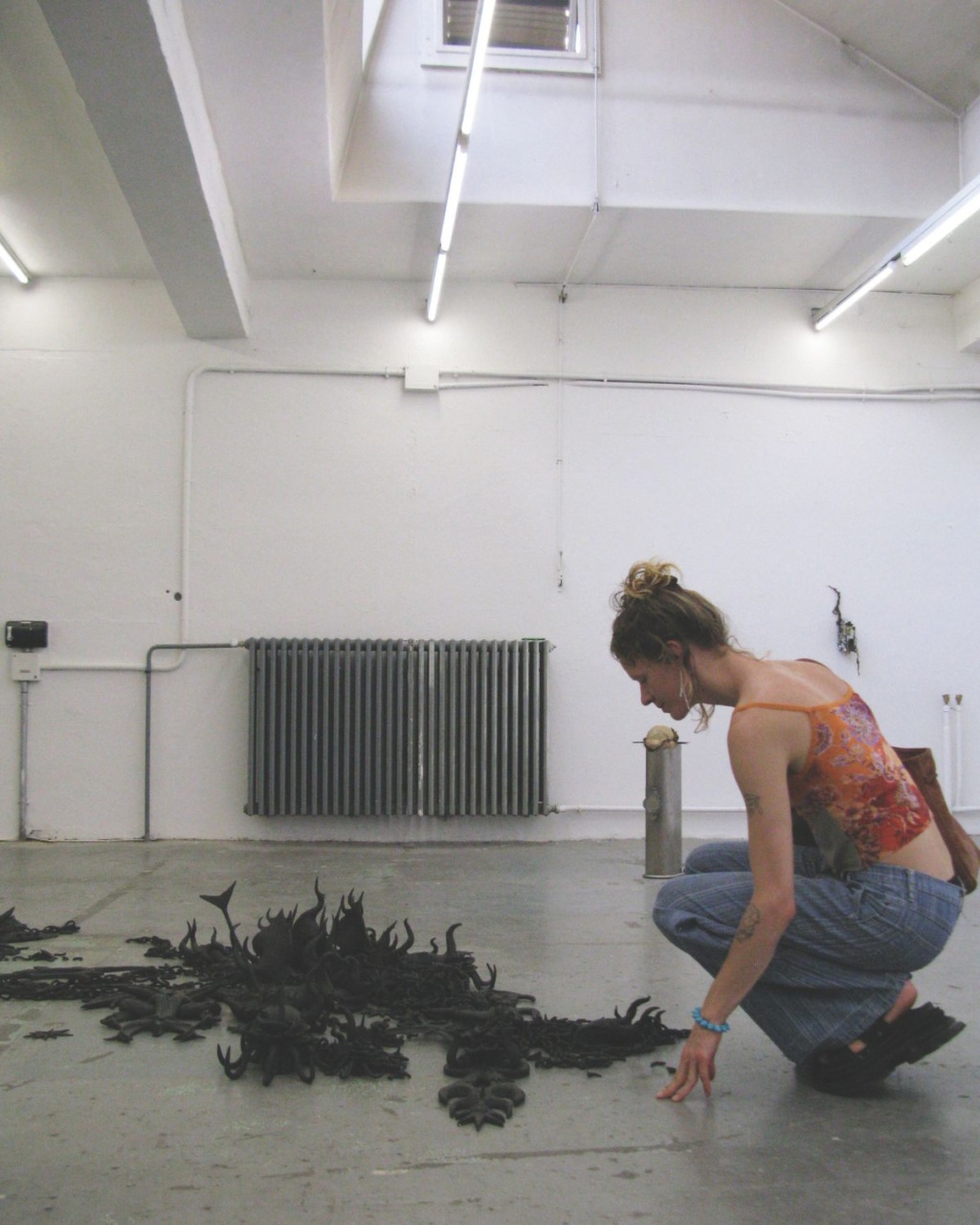 Luca crouches, curiously examining a cluster of dark, abstract sculptures on a concrete floor in an industrial-style room with fluorescent lighting.
