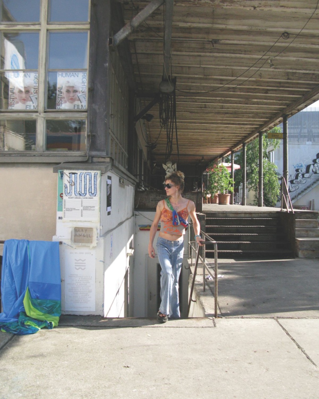 Luca stands near a building entrance, with large windows displaying posters. A covered walkway leads to stairs. Urban and casual vibe.