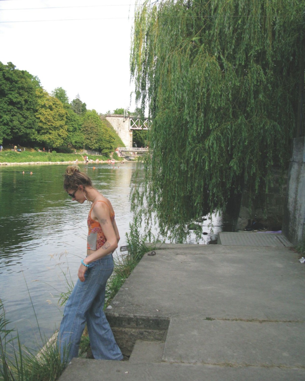 Luca stands by a riverbank, gazing down at the water, surrounded by lush greenery and a weeping willow.