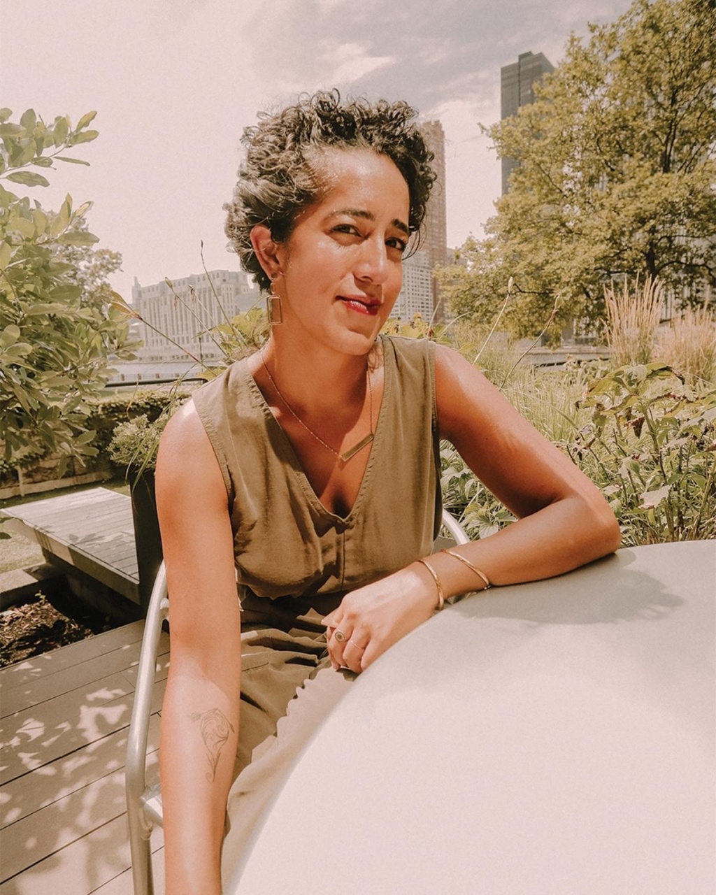 A woman with short curly hair sits at a round table outdoors on a sunny day. She wears a sleeveless dress and has a calm, relaxed expression.