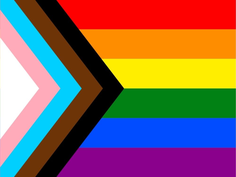Progress Pride Flag with horizontal rainbow stripes and a chevron on the left. Chevron includes white, pink, blue, brown, and black stripes, symbolizing inclusivity.